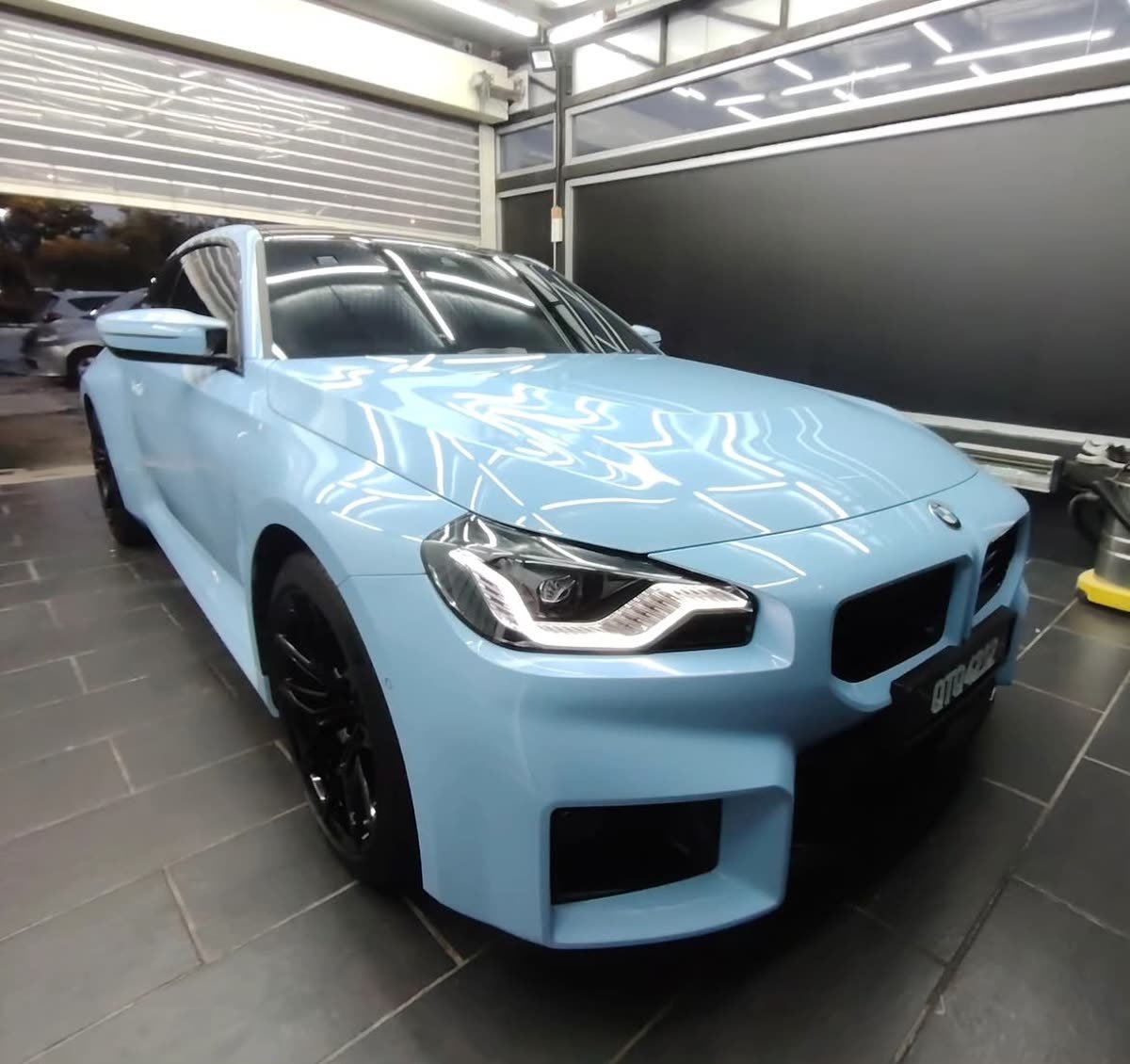 A powder-blue BMW M2 with paint protection film installed, front three-quarter view in a different garage setting