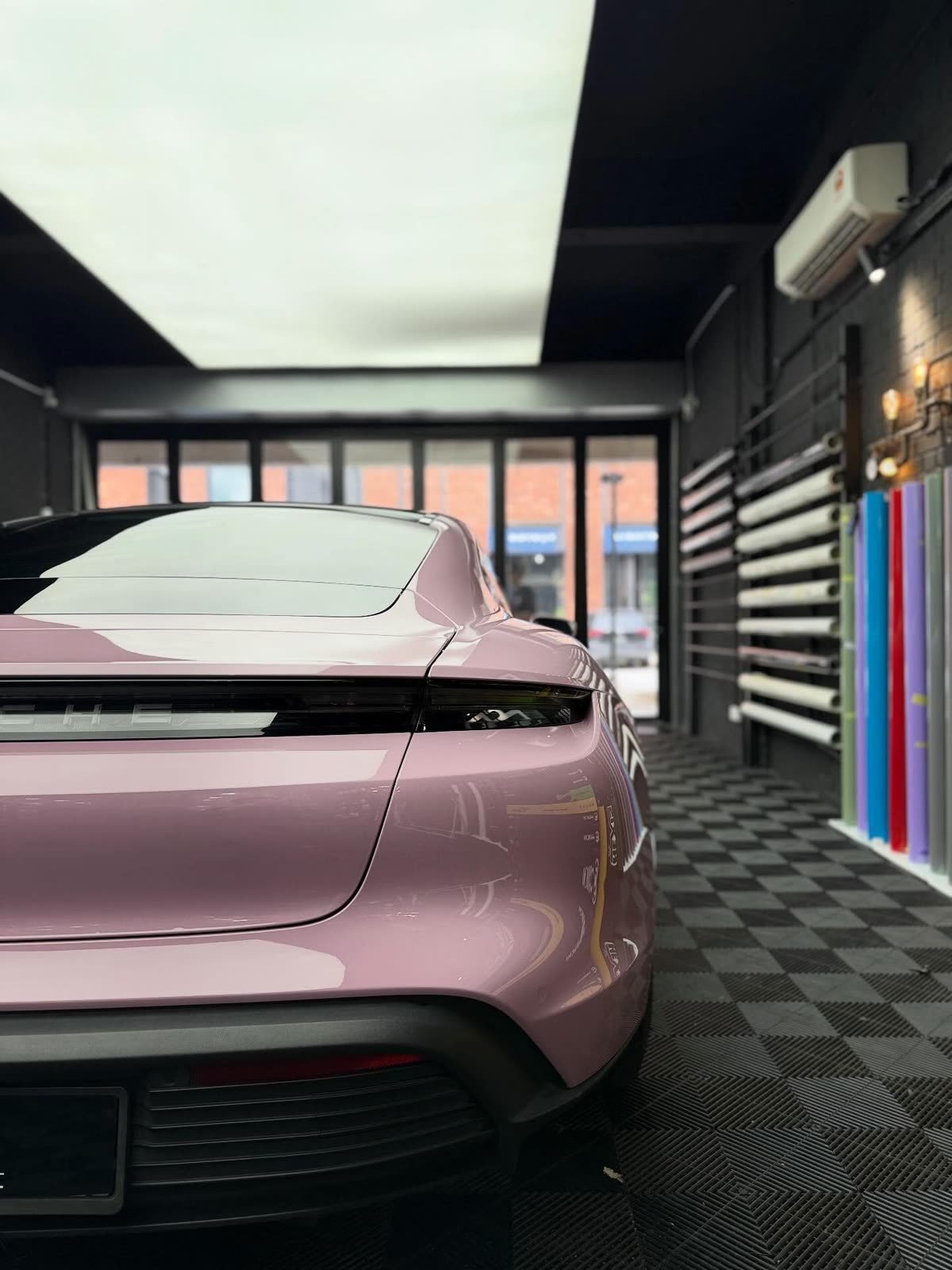A pink Porsche Taycan, rear view beside a rack of colour vinyl rolls