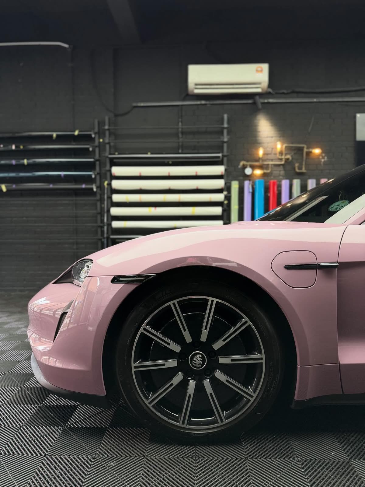 A pink Porsche Taycan, close-up of the front wheel and fender beside a rack of vinyl rolls