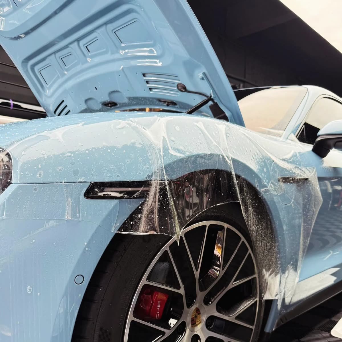 Close-up of paint protection film being applied over a powder-blue Porsche Taycan's front fender and headlight, soapy solution visible
