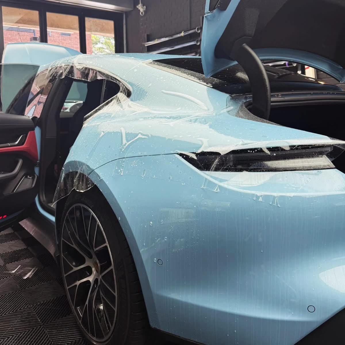 Paint protection film being smoothed over a powder-blue Porsche Taycan's rear quarter panel, trunk open