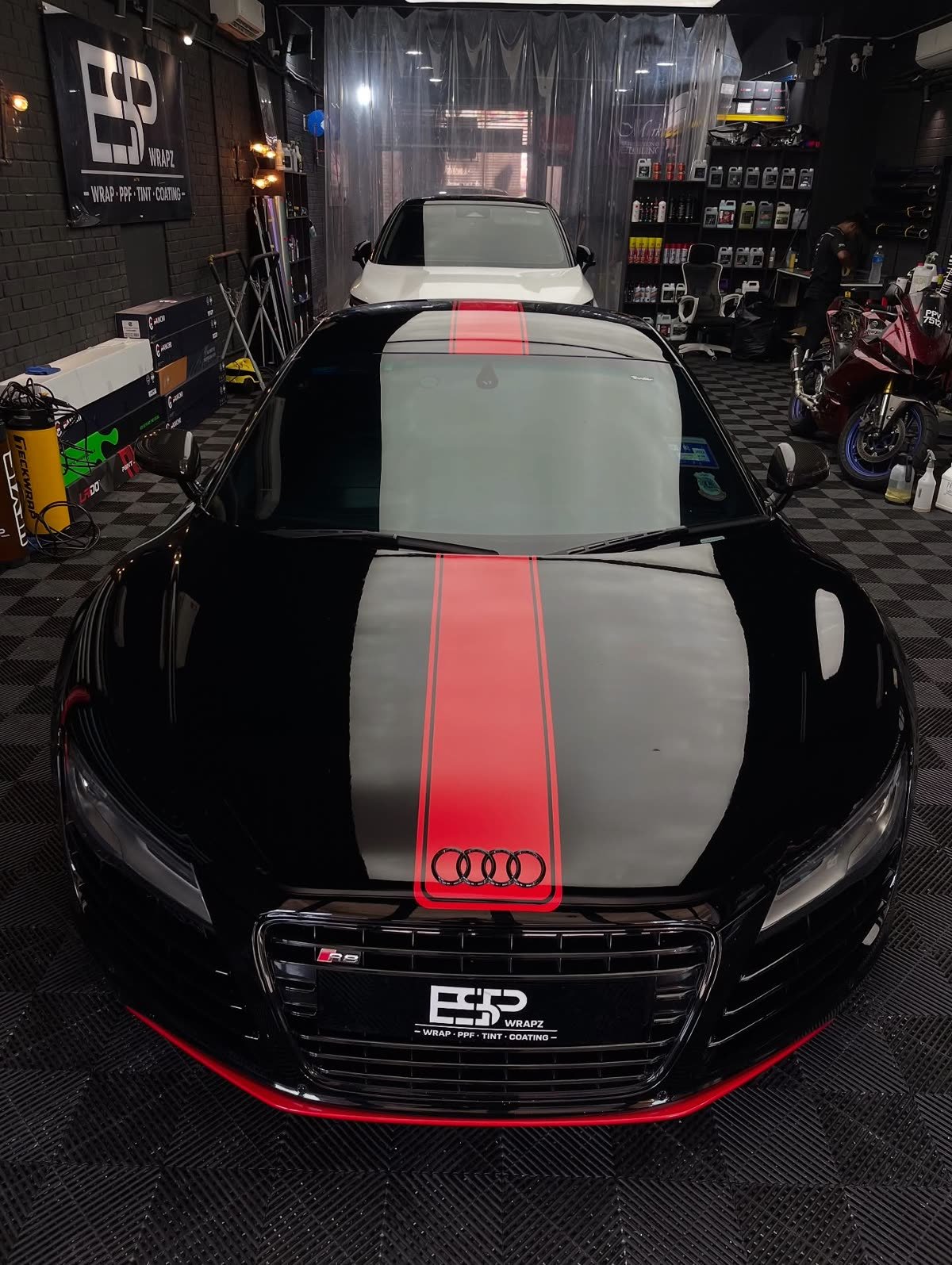 A black Audi R8 with a red racing stripe wrap, front view of the workshop with motorcycles parked nearby