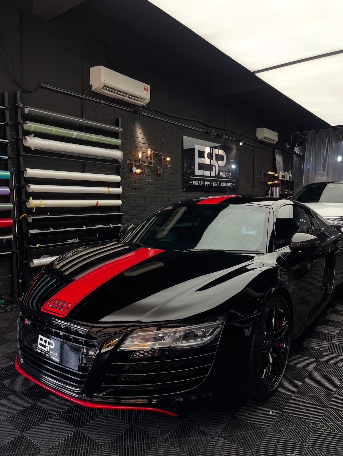 A black Audi R8 with a red racing stripe wrap, front three-quarter view beside the vinyl roll rack and ESP Wrapz sign