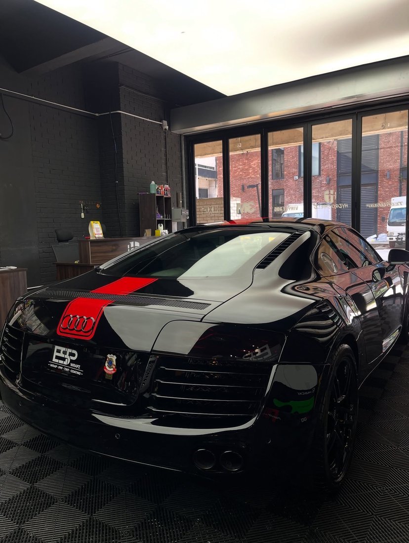 A black Audi R8 with a red racing stripe wrap, front view inside the workshop with colour vinyl rolls on the wall