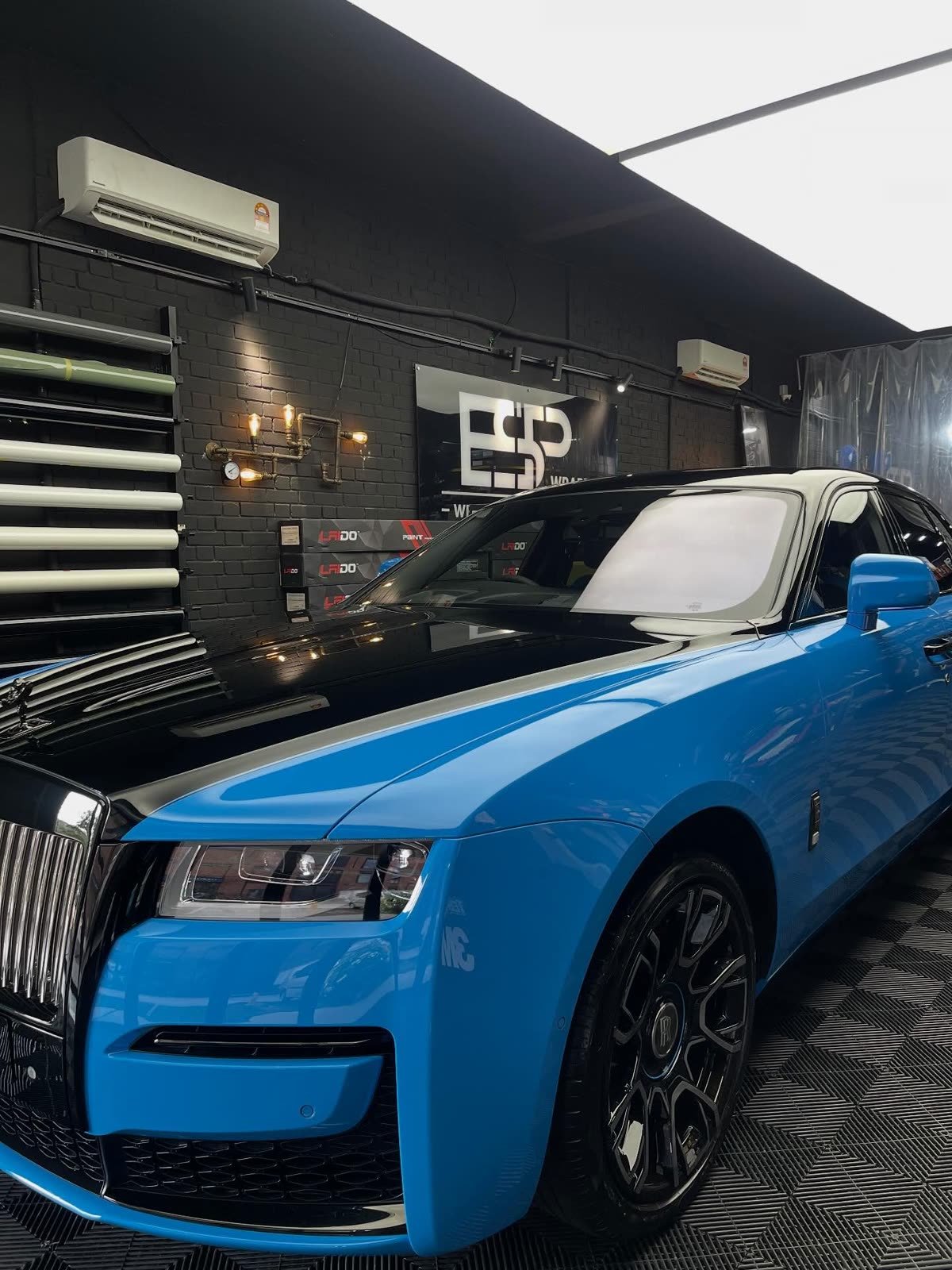 A Rolls-Royce Ghost with a bright blue and black two-tone colour wrap, front three-quarter view beside boxes of PPF film