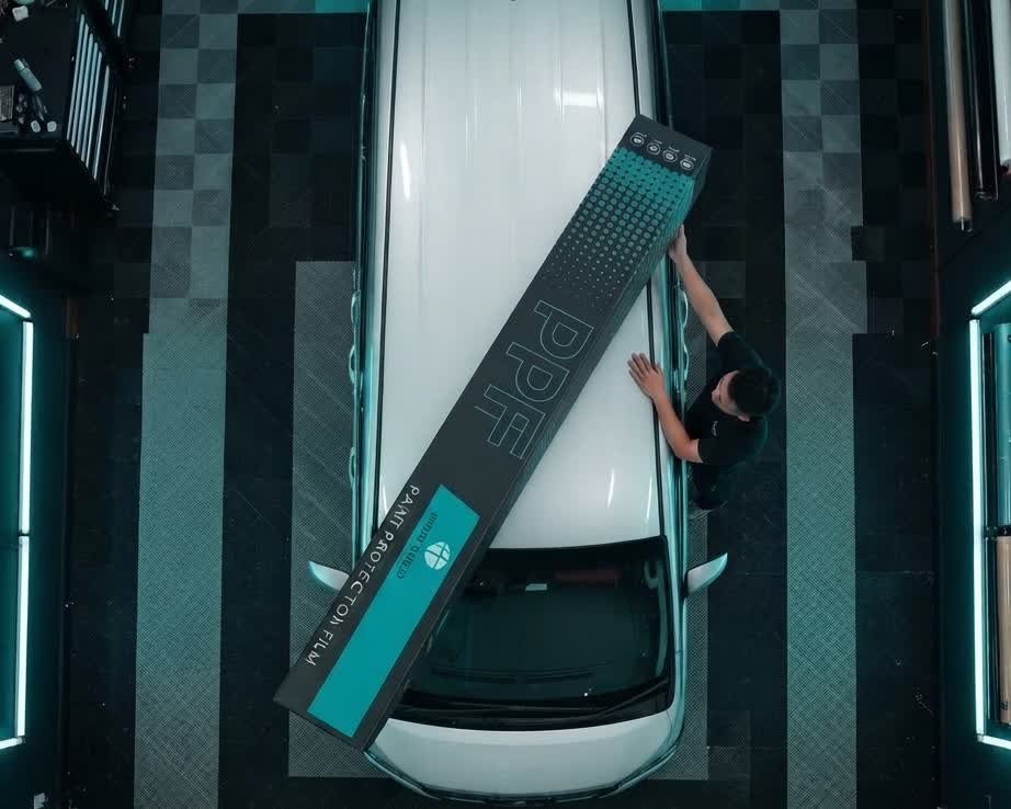 Aerial view of a technician placing a Hydroshield PPF box on a vehicle roof in a detailing workshop, rendered illustration